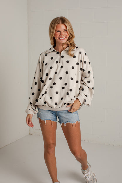 A woman modeling a cream-colored half-zip hoodie with black polka dots, paired with denim shorts and white sneakers, standing against a white brick wall.