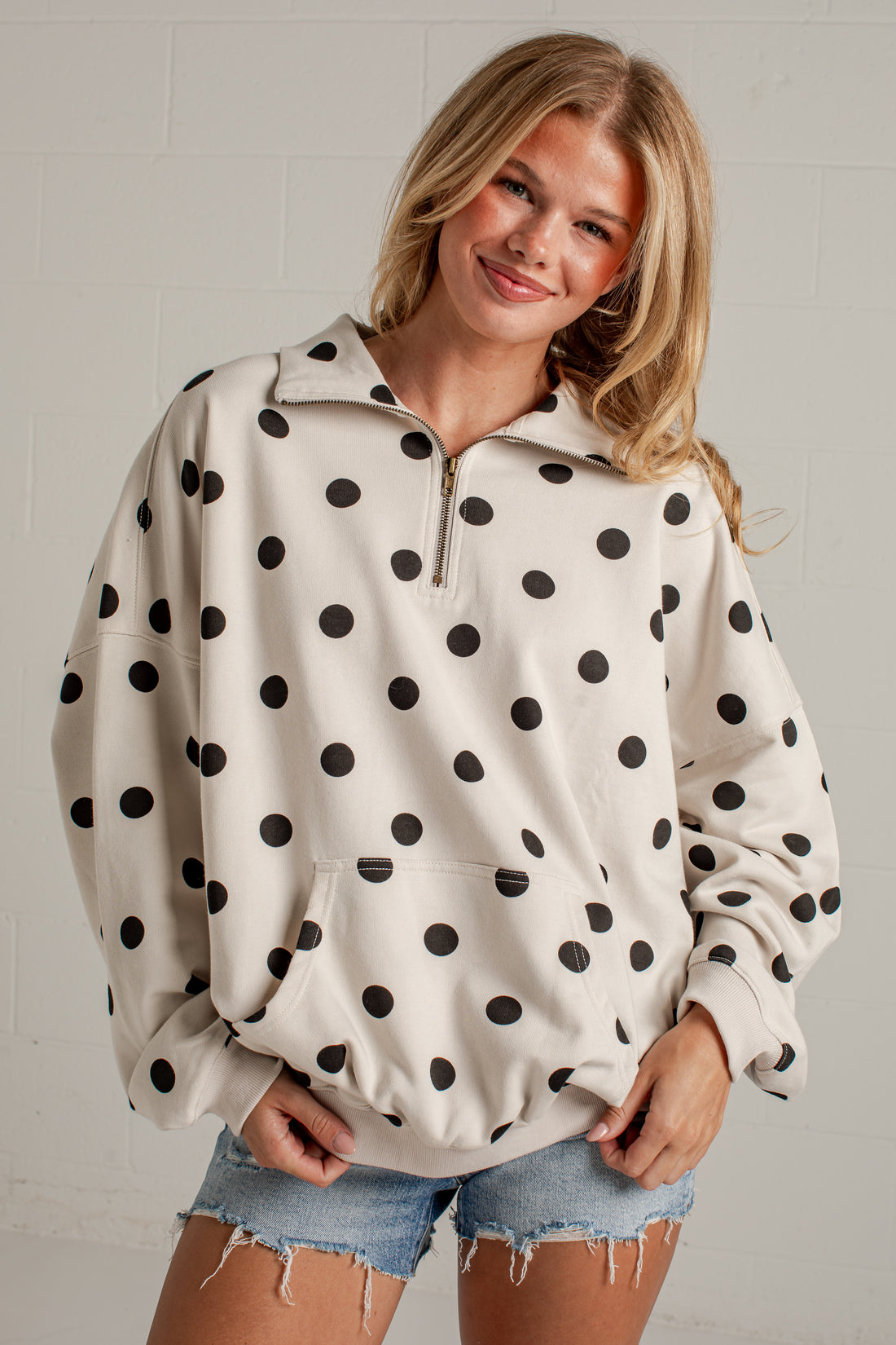 A woman wearing a cream-colored half-zip sweatshirt with black polka dots and denim shorts, standing against a light gray brick wall.