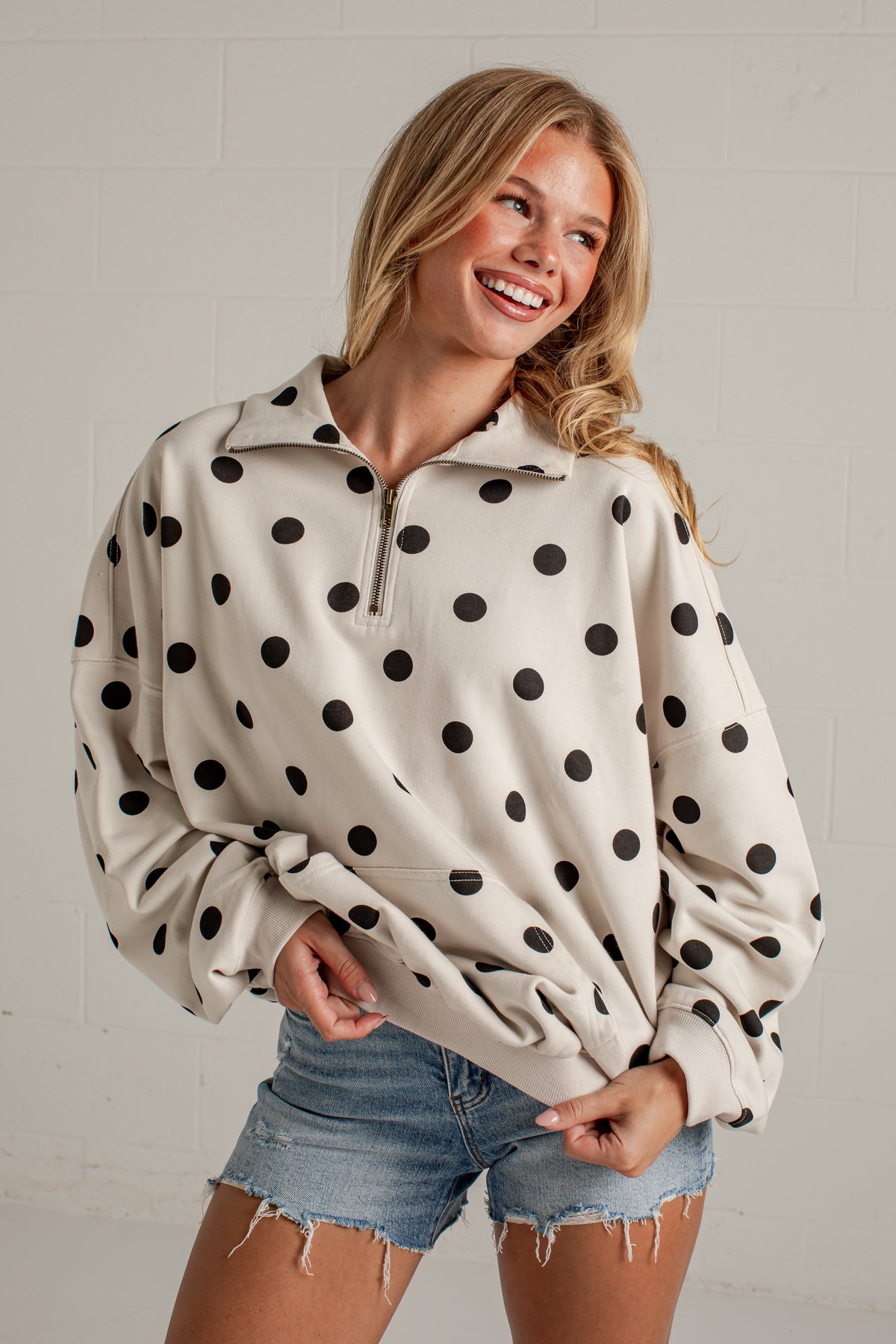 A woman wearing a cream-colored half-zip sweatshirt with black polka dots, paired with denim shorts, posing against a white brick wall.