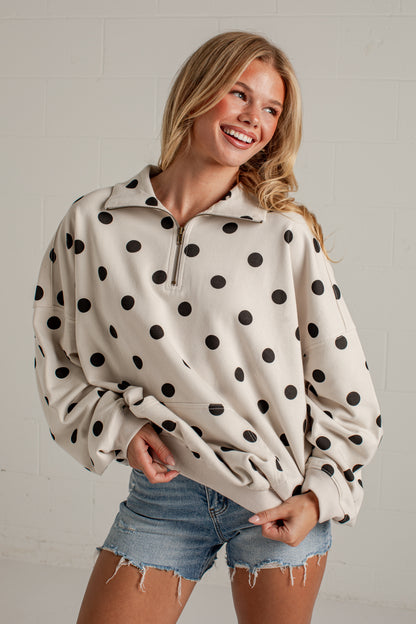 A woman wearing a cream-colored half-zip sweatshirt with black polka dots, paired with denim shorts, posing against a white brick wall.