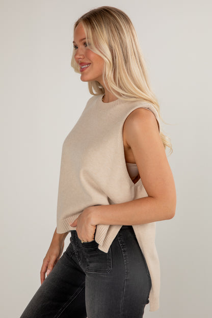 Woman wearing a beige sleeveless top and dark jeans against a plain background