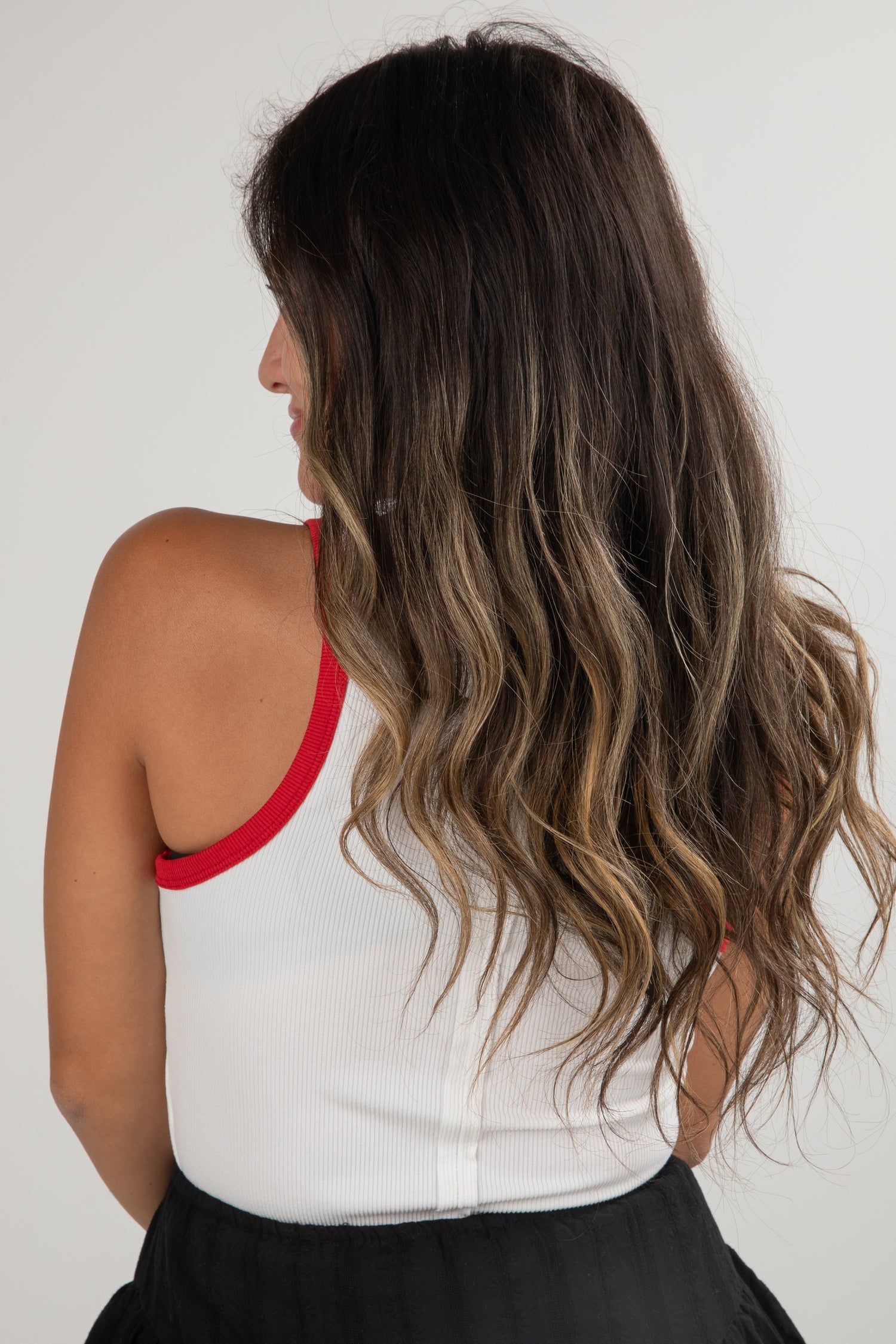 Woman with long, wavy hair wearing a white top with red trim and black pants on a plain background