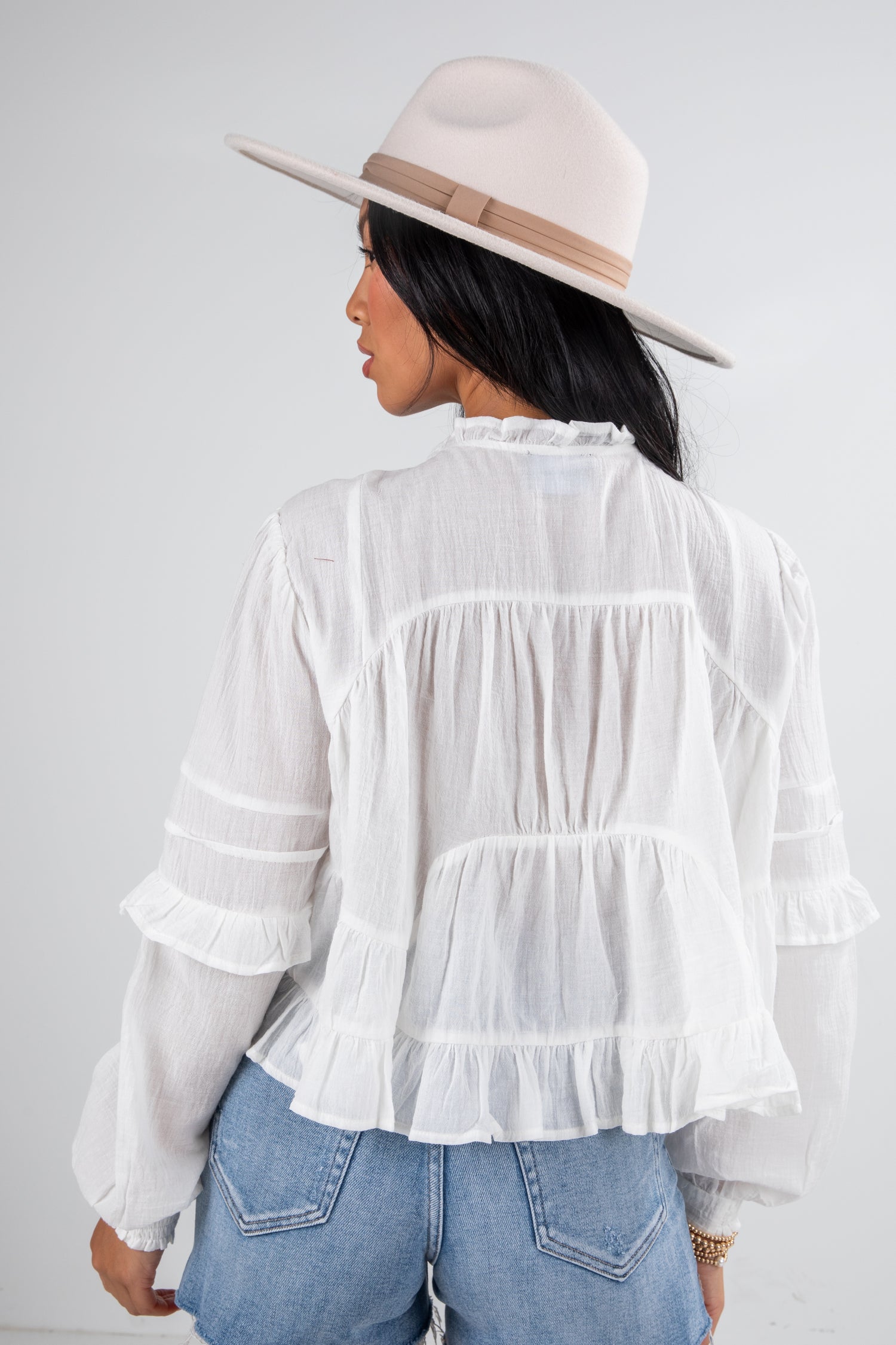 Person wearing a white ruffled blouse and blue jeans with a beige hat on a plain background