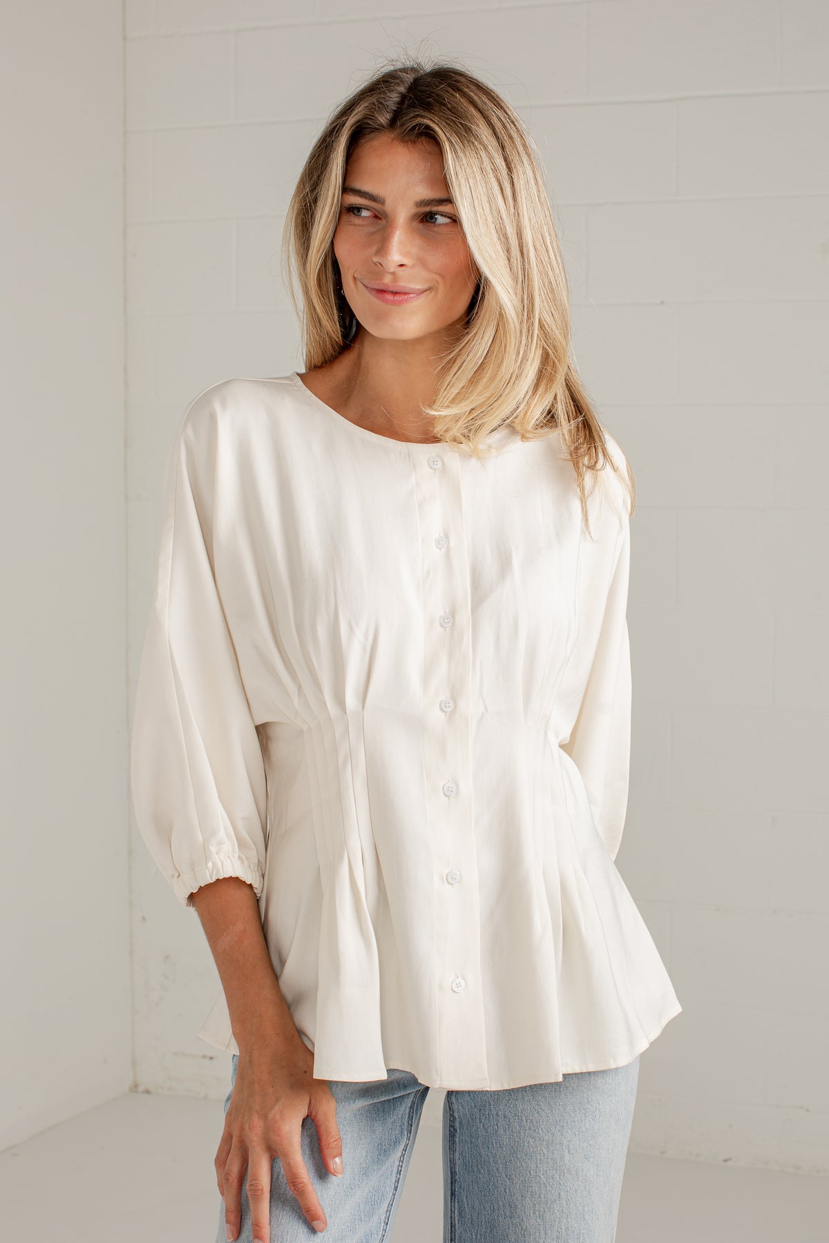 A woman wearing a cream-colored, long-sleeved button-up blouse with a gathered peplum waist, paired with light blue jeans, standing against a white brick wall.