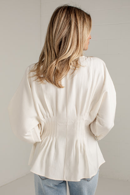 A woman viewed from behind wearing a cream-colored long-sleeve blouse with a gathered peplum waist, paired with light blue jeans, set against a neutral light gray background.