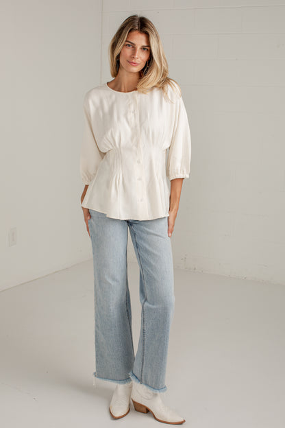 A woman modeling a cream-colored, long-sleeved peplum blouse with light-wash, wide-leg denim jeans and white ankle boots, standing in a minimalist white studio setting.