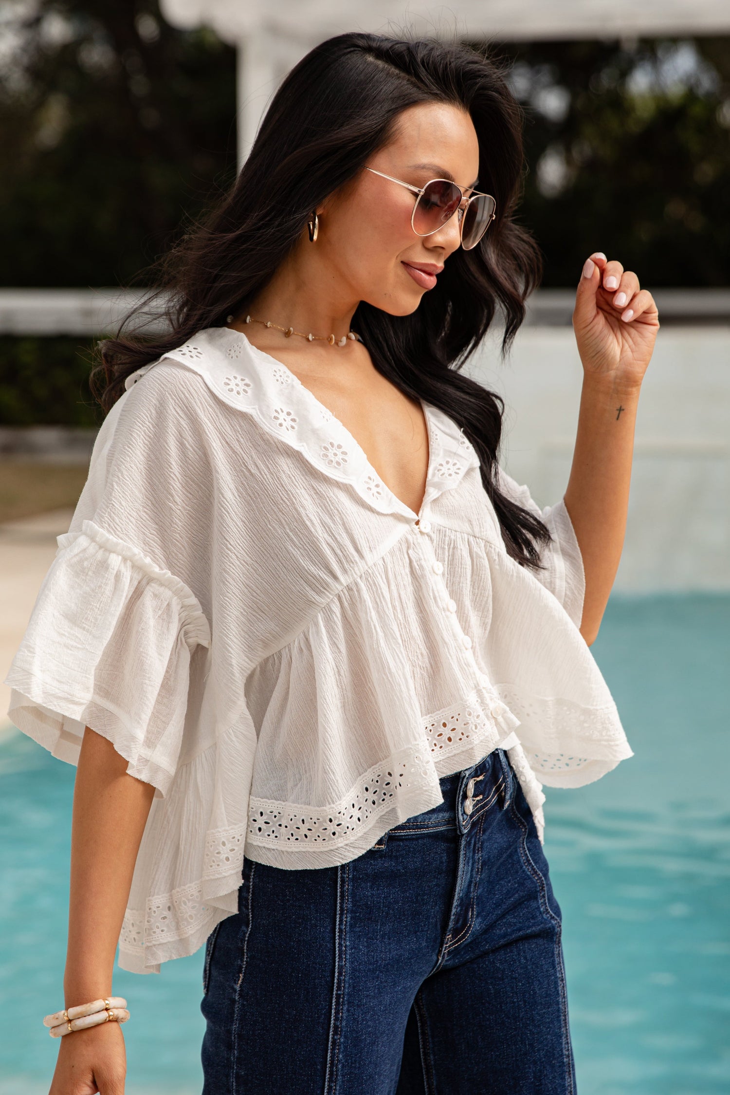Woman wearing a white blouse with ruffled sleeves and blue jeans by a pool.