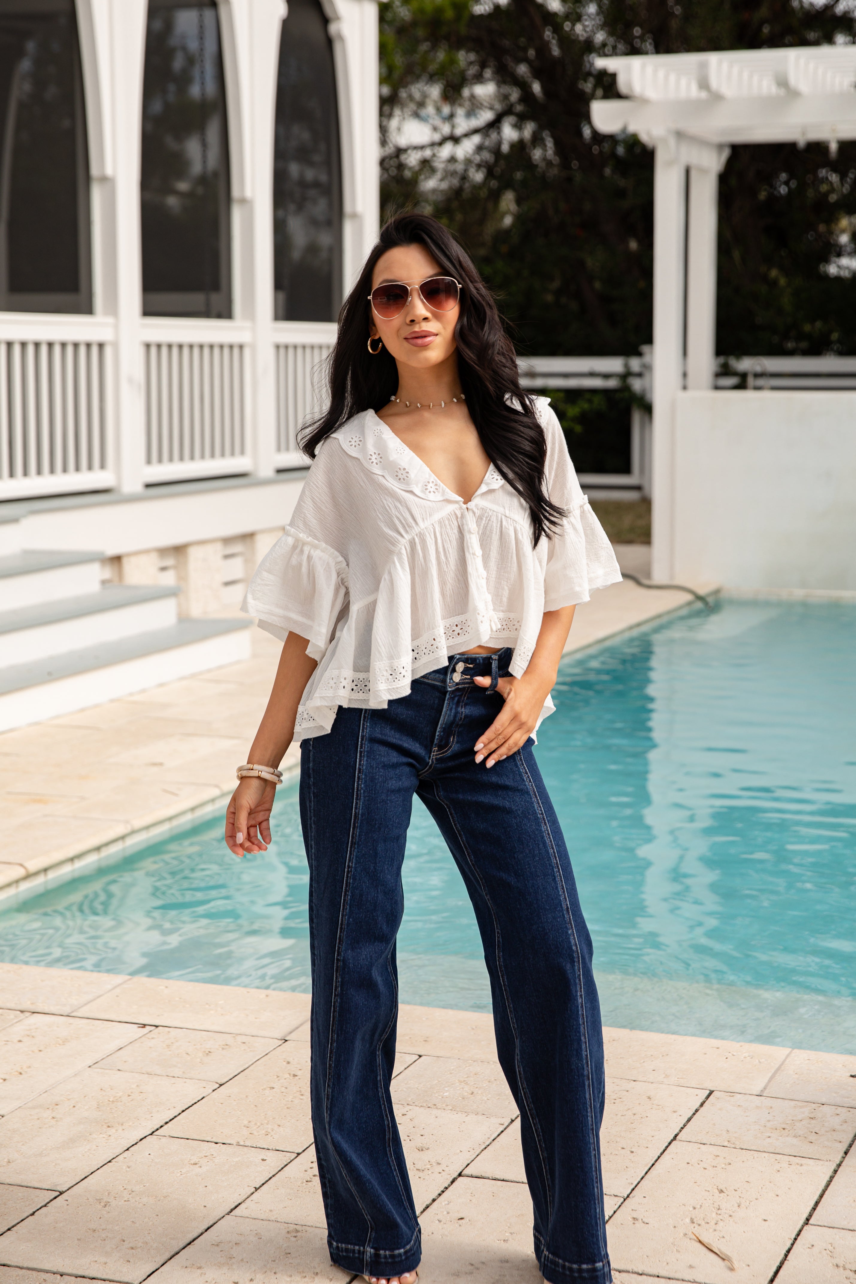 Woman in a white blouse and blue jeans standing by a pool.
