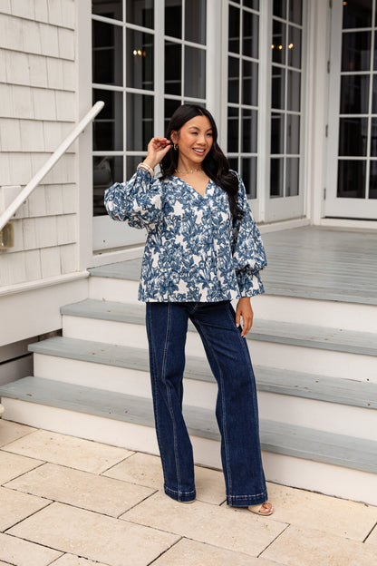 Woman wearing a blue floral blouse and jeans standing on a porch.