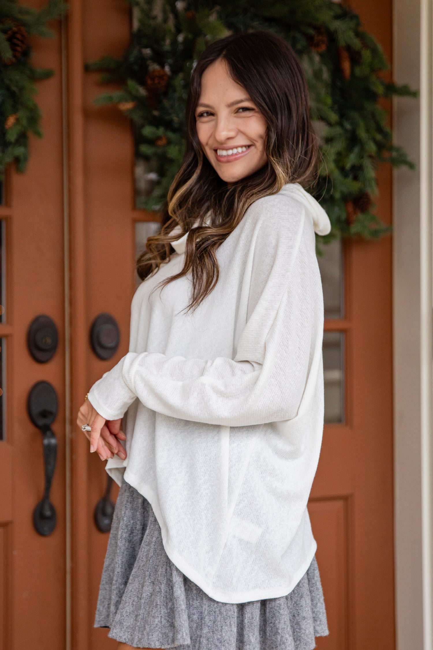 Woman wearing a light gray long-sleeve top and gray skirt standing in front of a wooden door with greenery.