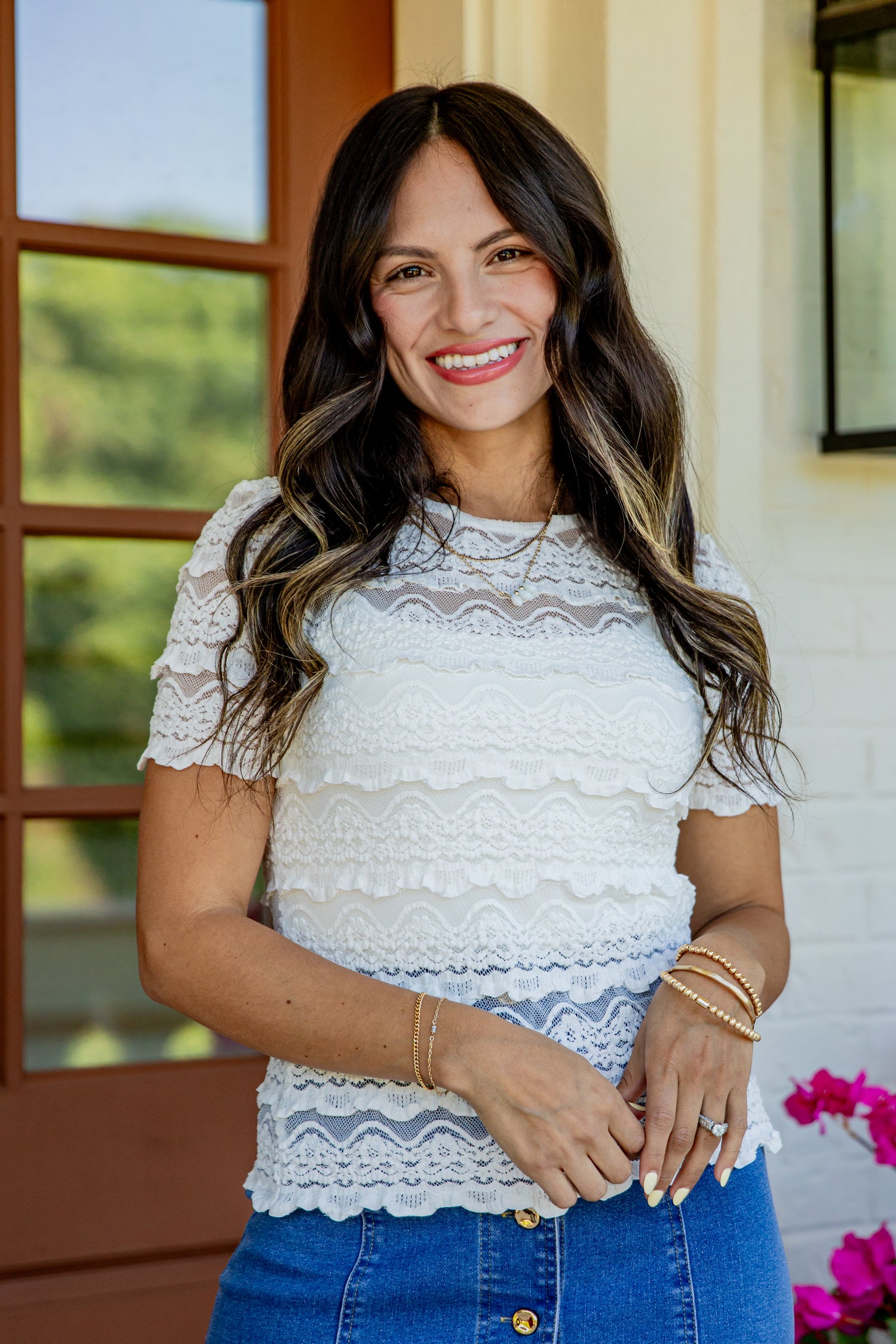 A woman smiling and wearing a white short-sleeve lace top with ruffle details, paired with blue jeans, standing in front of a porch with a brown door and white brick wall.