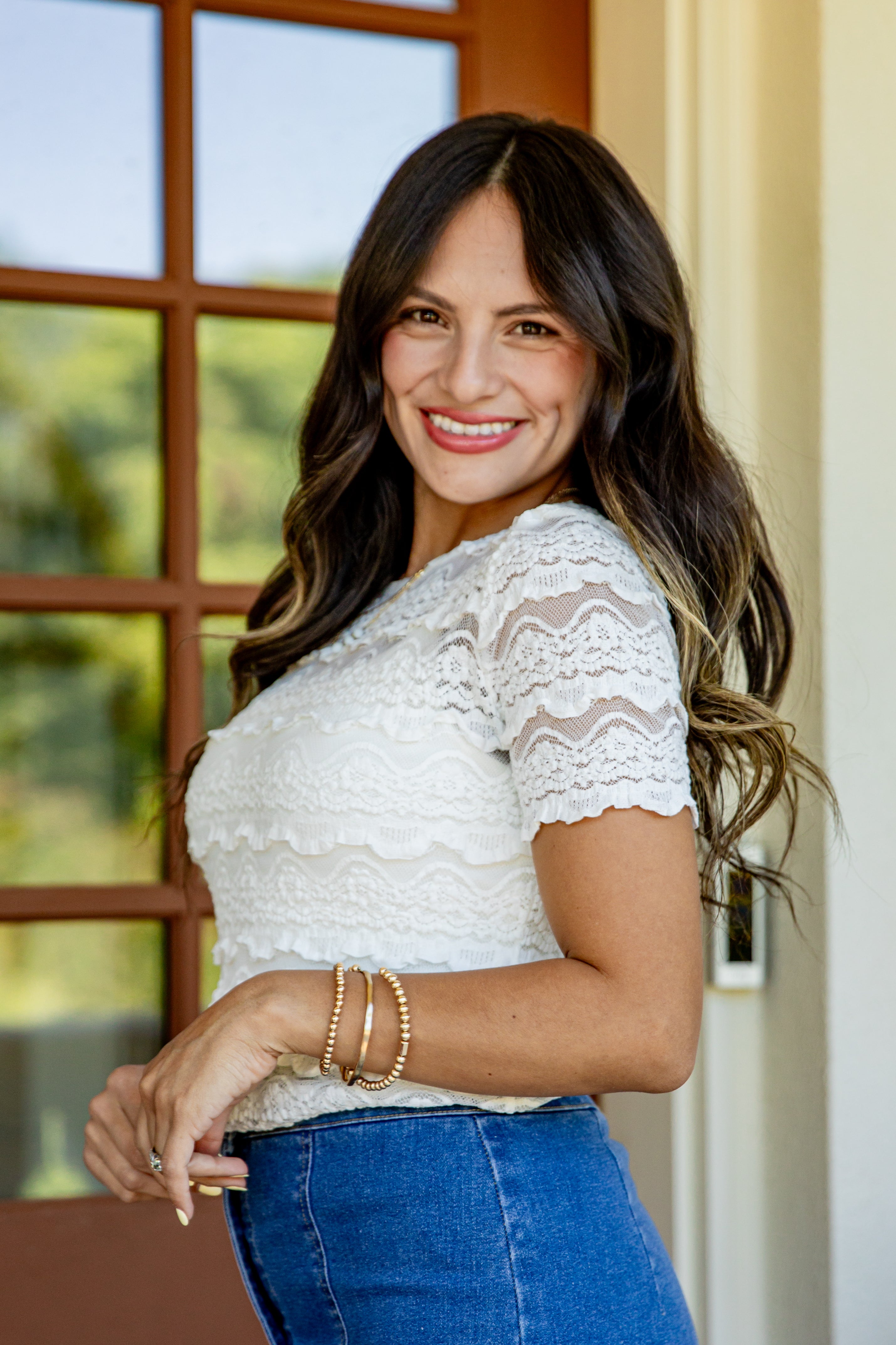 A smiling woman with long dark brown hair wearing a white lace short-sleeve top and blue jeans, standing indoors in front of a window.