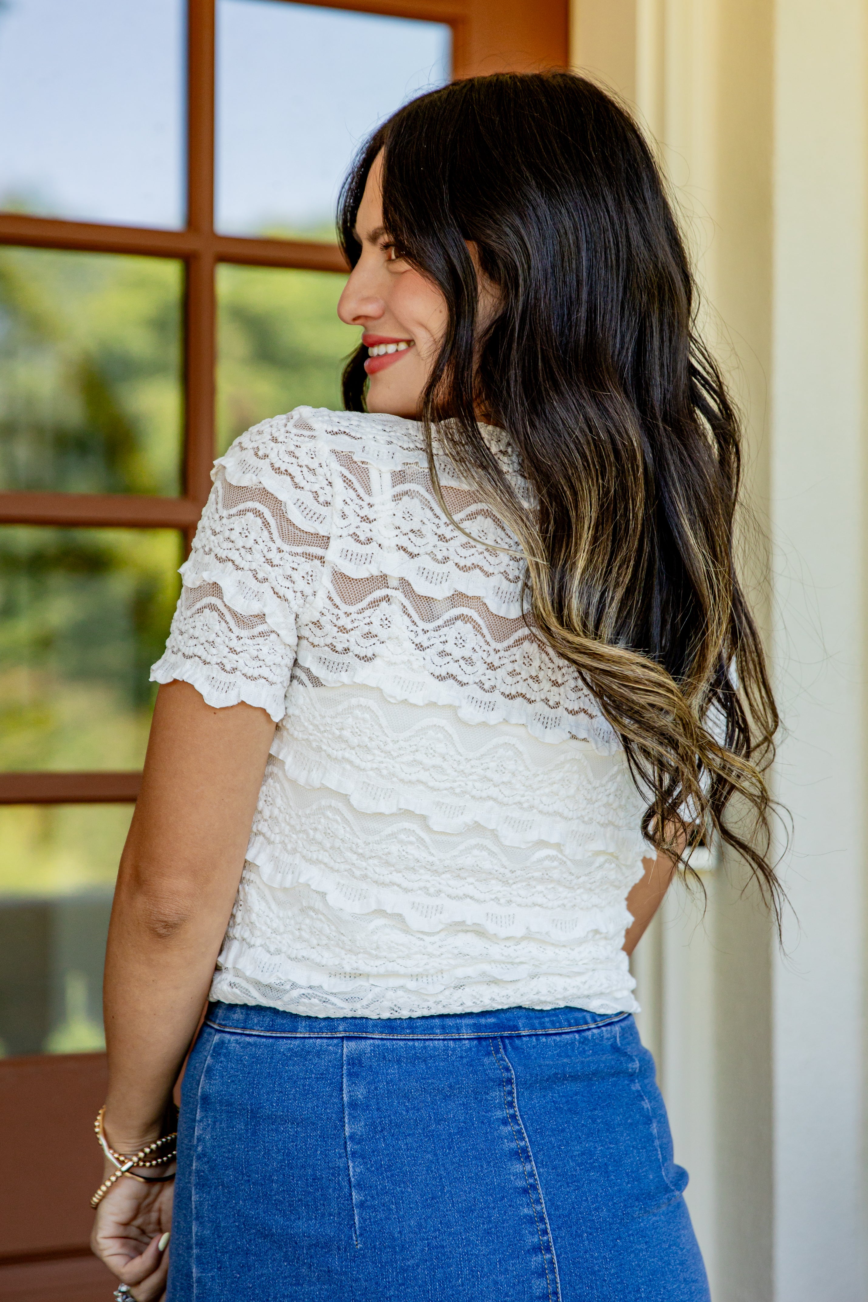 A woman with long dark brown hair with blonde highlights is shown from the back wearing a white lace short-sleeve top and a blue denim skirt, standing indoors near a window.