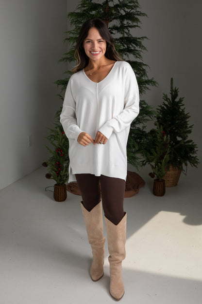Woman wearing a white long-sleeve top and brown pants with beige boots, standing in front of decorative plants.
