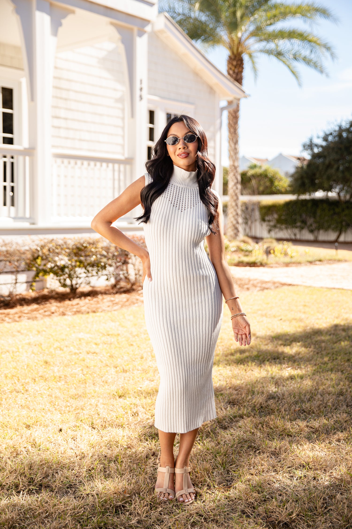 Woman in a white ribbed dress standing outdoors with a house and palm tree in the background.