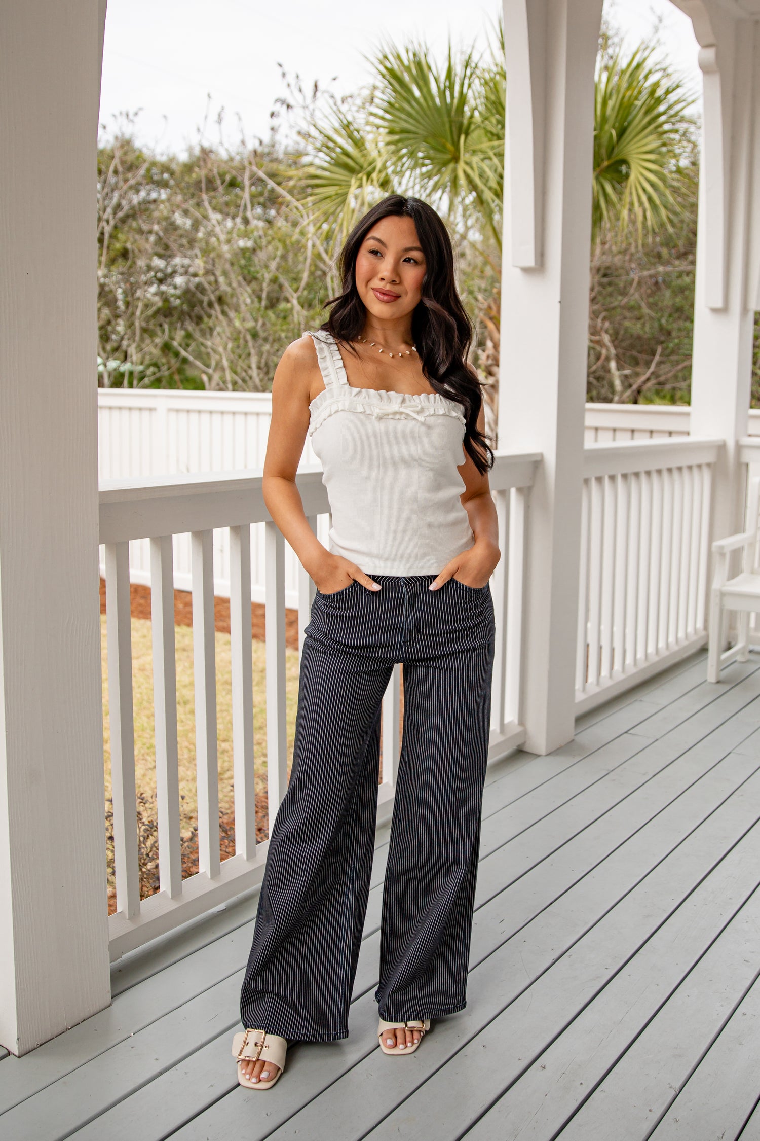 Woman standing on a porch wearing a white top and dark pants.