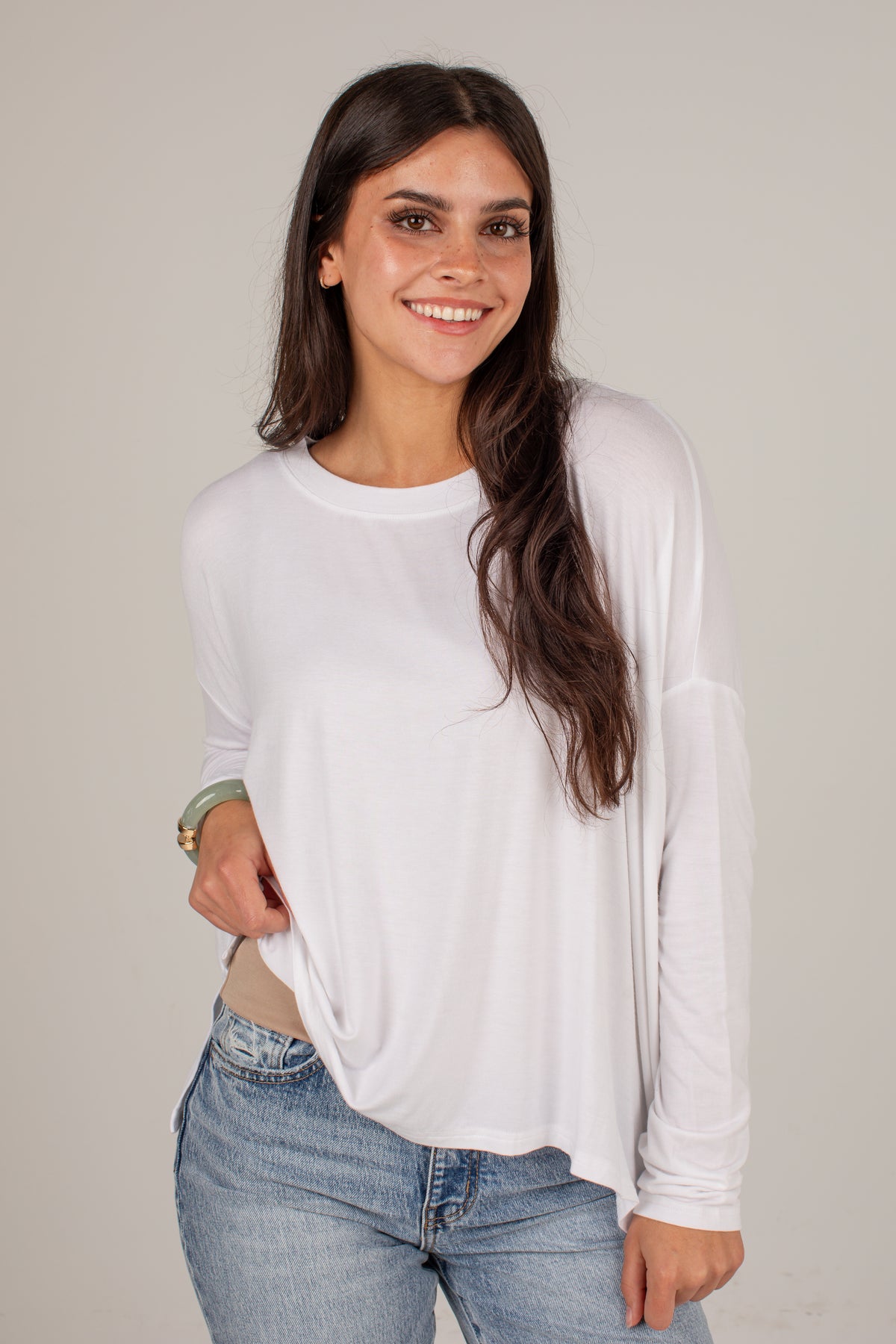 A woman with long dark hair smiling, wearing a white long-sleeve crewneck top and light blue jeans, standing against a plain light gray studio background.