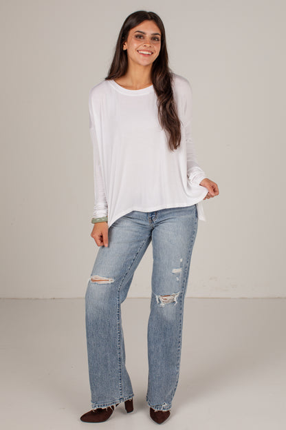 A woman modeling a white long-sleeve top and light-wash distressed jeans with ripped knees, standing against a plain light beige studio background.
