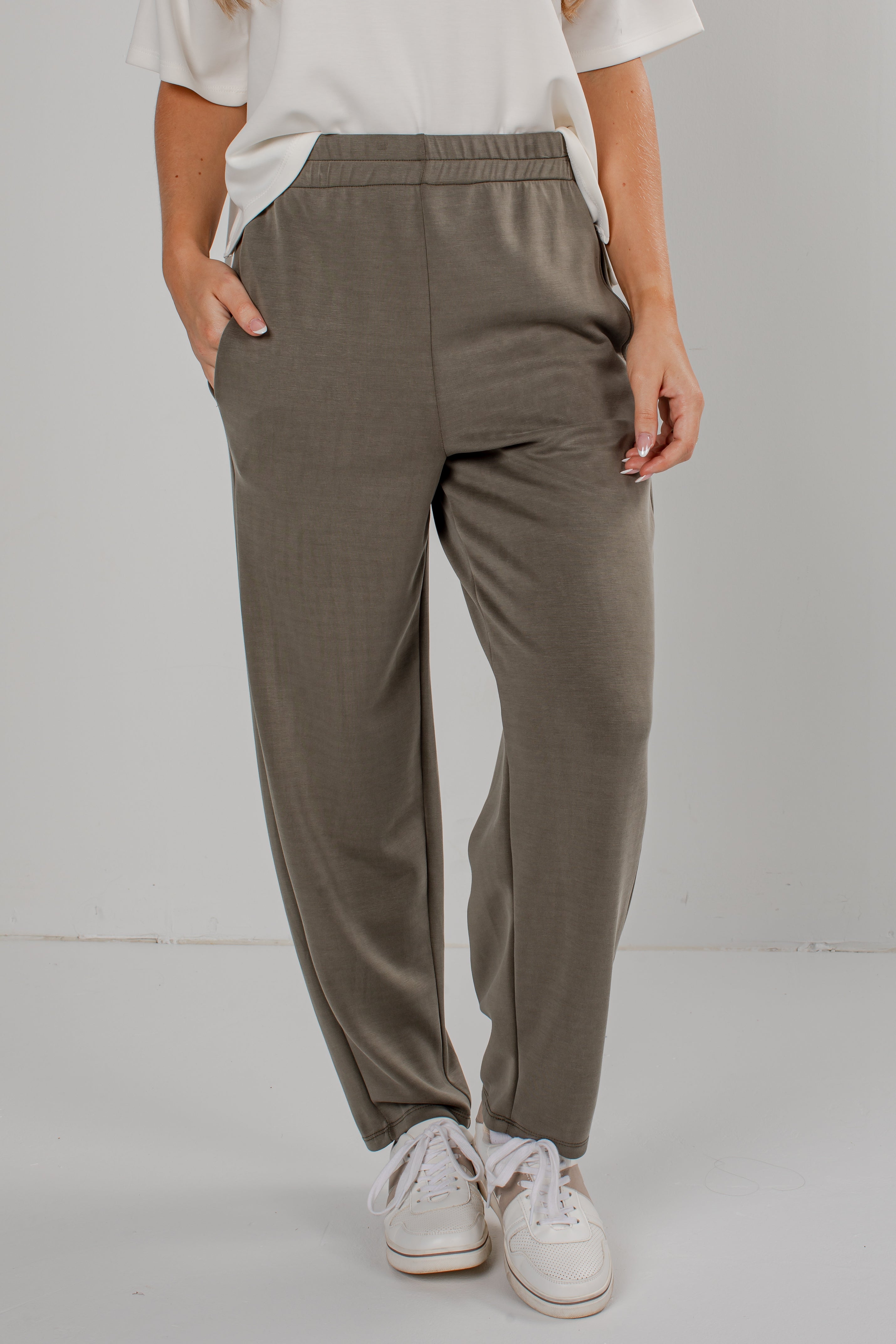 A person wearing loose, straight-leg jogger pants in a muted taupe-gray color, paired with a white t-shirt and white sneakers, standing against a light gray studio background.
