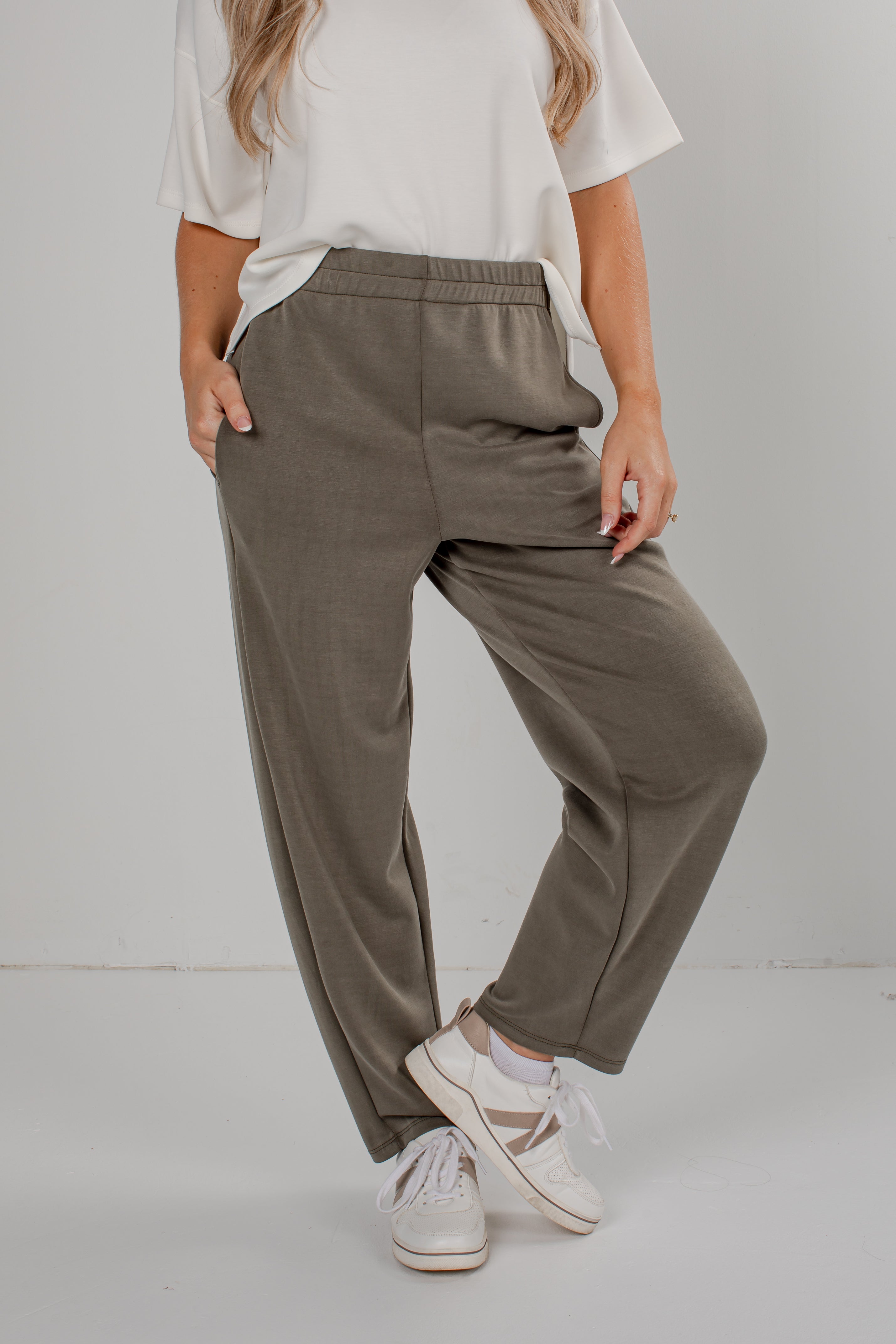 A person wearing loose, wide-leg olive green pants with an elastic waistband, styled with a white t-shirt and white sneakers against a plain light gray studio background.