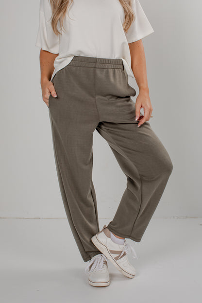 A person wearing loose, wide-leg olive green pants with an elastic waistband, styled with a white t-shirt and white sneakers against a plain light gray studio background.