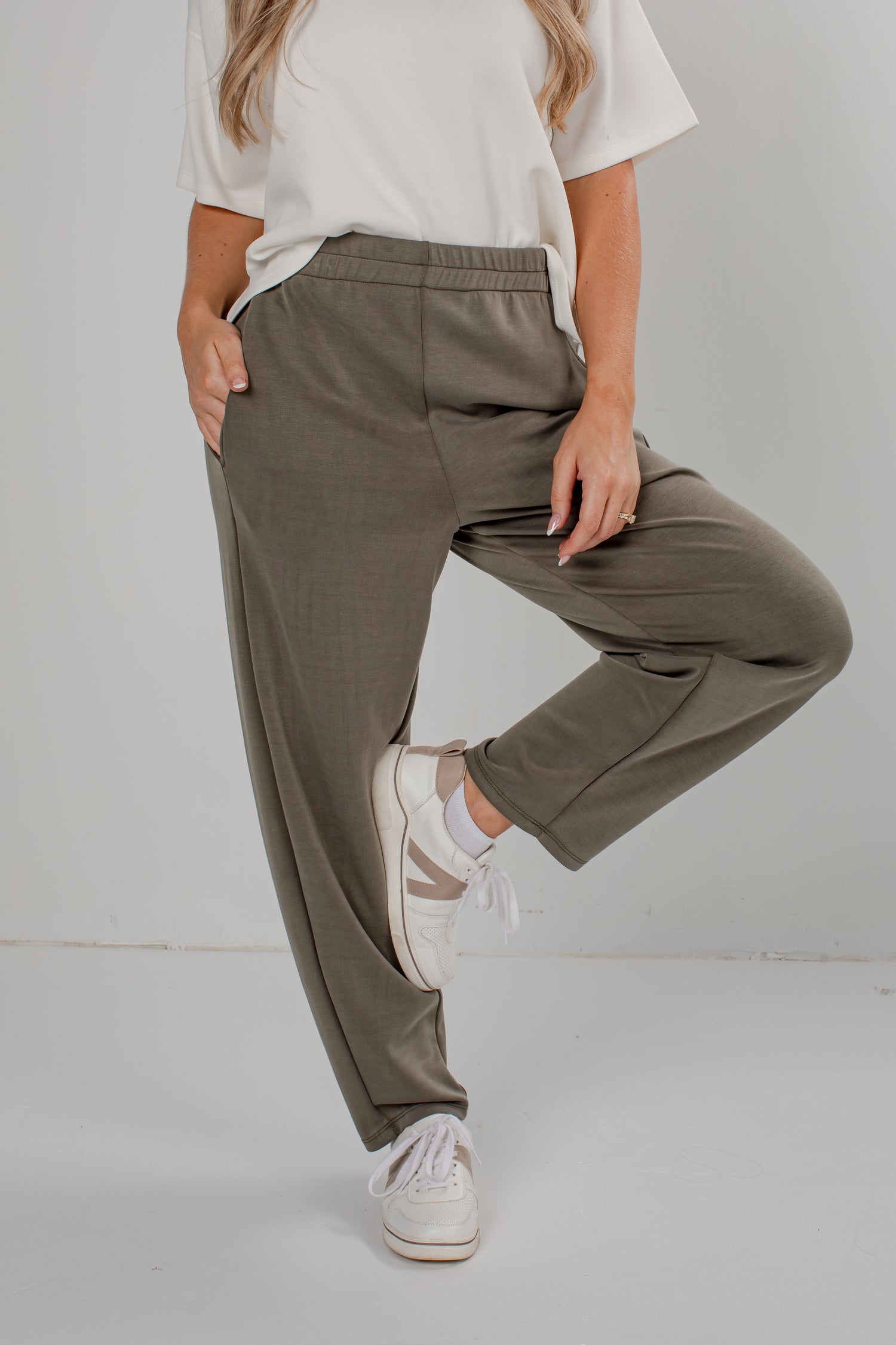 A person wearing loose, straight-leg olive green pants with a white t-shirt and white sneakers, standing against a light gray studio background.