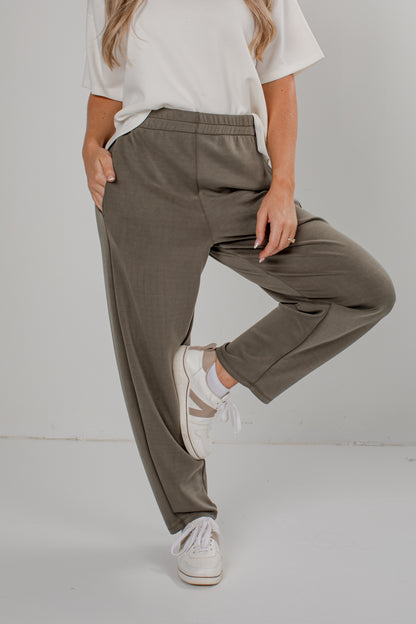 A person wearing loose, straight-leg olive green pants with a white t-shirt and white sneakers, standing against a light gray studio background.