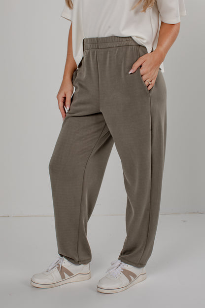 A person wearing loose, straight-leg jogger-style pants in a muted taupe/olive-gray color, paired with a white top and white sneakers, standing against a light gray studio background.