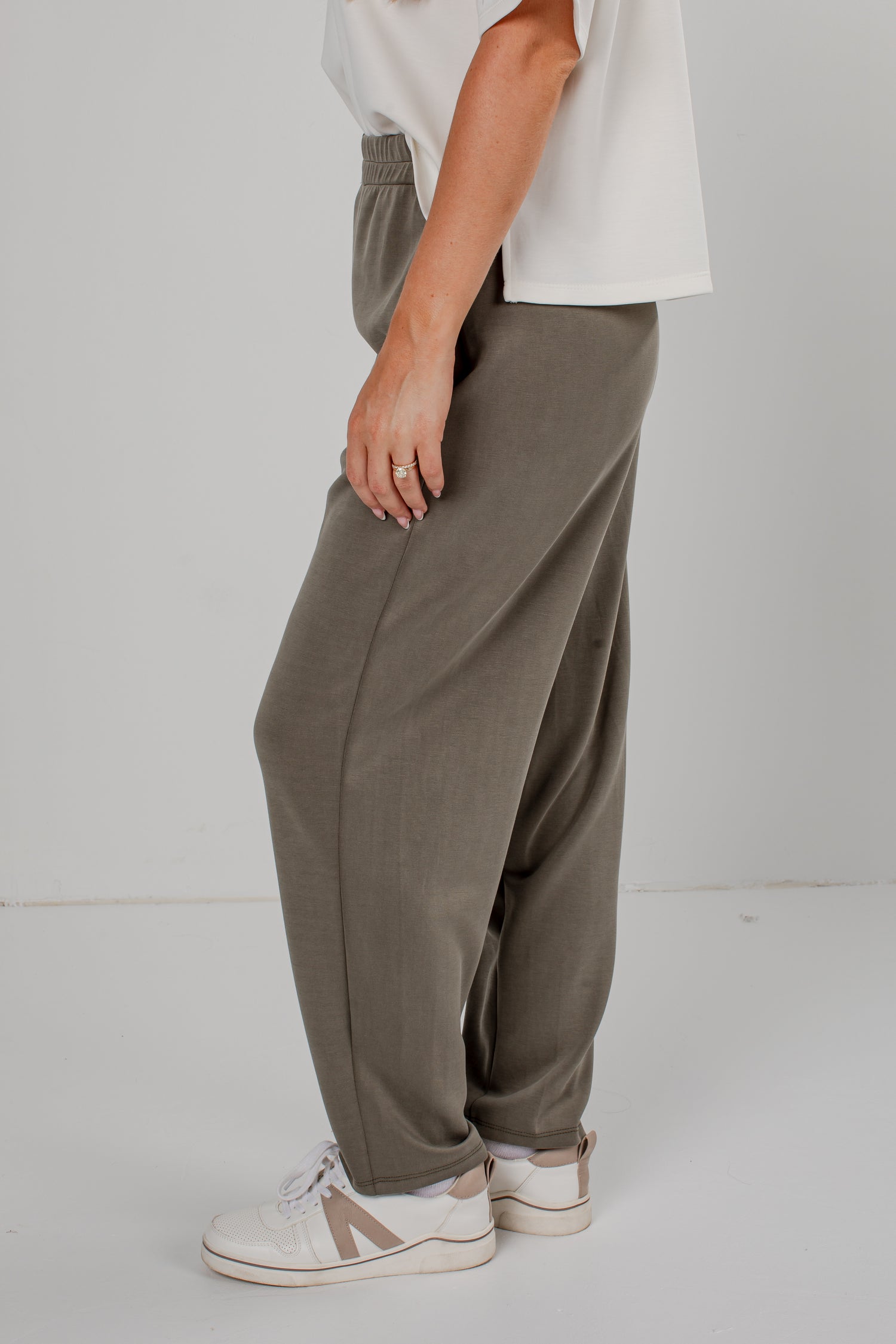 A person wearing loose, wide-leg taupe pants with an elastic waistband, paired with a white t-shirt and white sneakers, shown from the waist down against a light gray studio background.