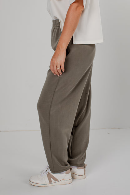 A person wearing loose, wide-leg taupe pants with an elastic waistband, paired with a white t-shirt and white sneakers, shown from the waist down against a light gray studio background.