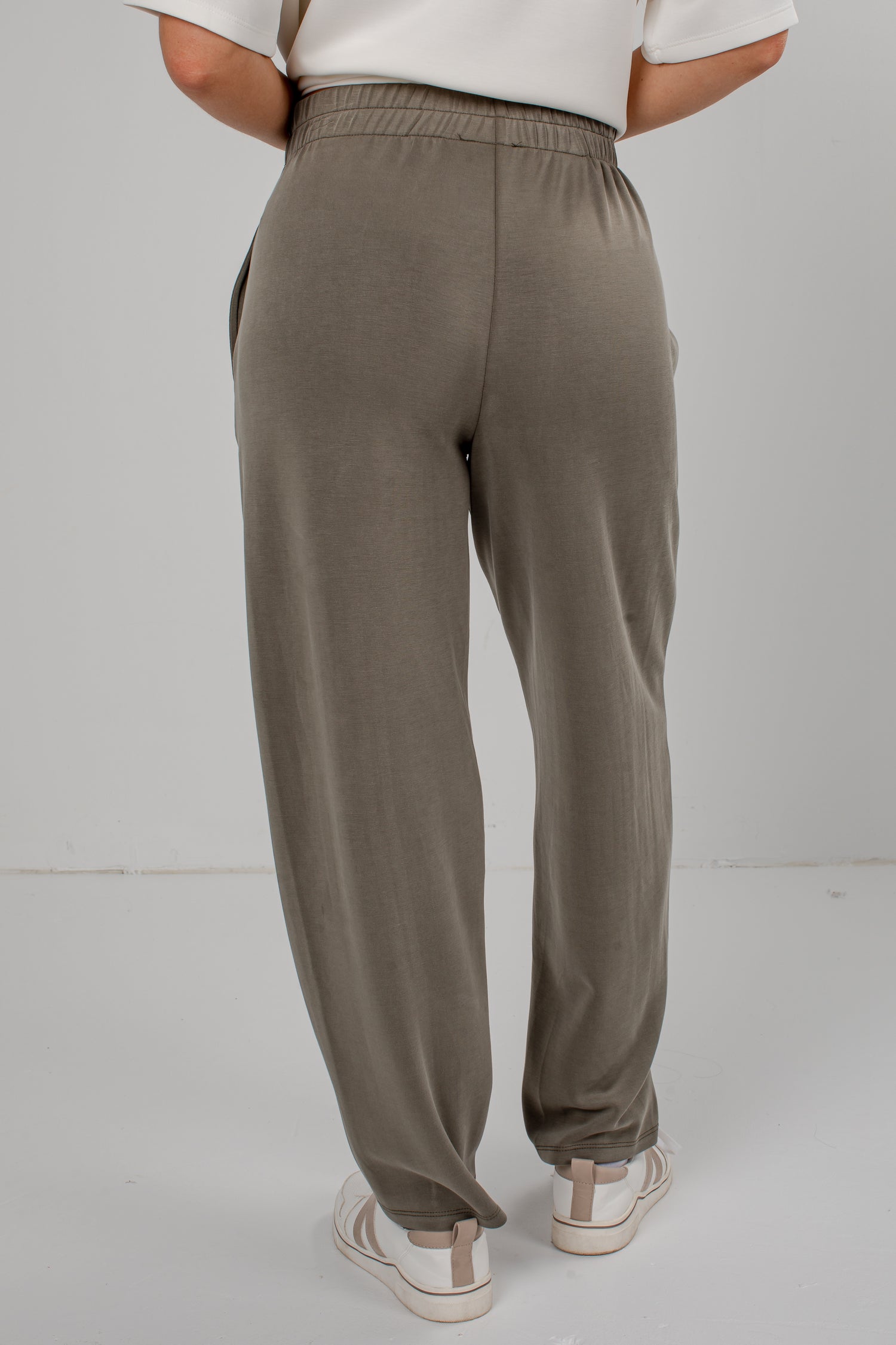 A person shown from the back wearing loose, wide-leg jogger-style pants in a muted taupe/olive-brown tone, paired with a white top and white sneakers, against a plain light gray studio background.