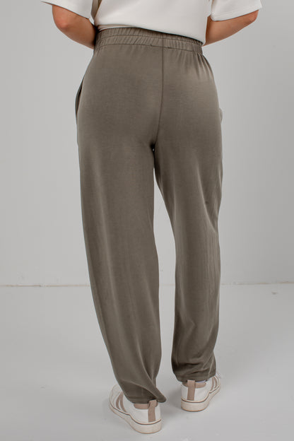 A person shown from the back wearing loose, wide-leg jogger-style pants in a muted taupe/olive-brown tone, paired with a white top and white sneakers, against a plain light gray studio background.