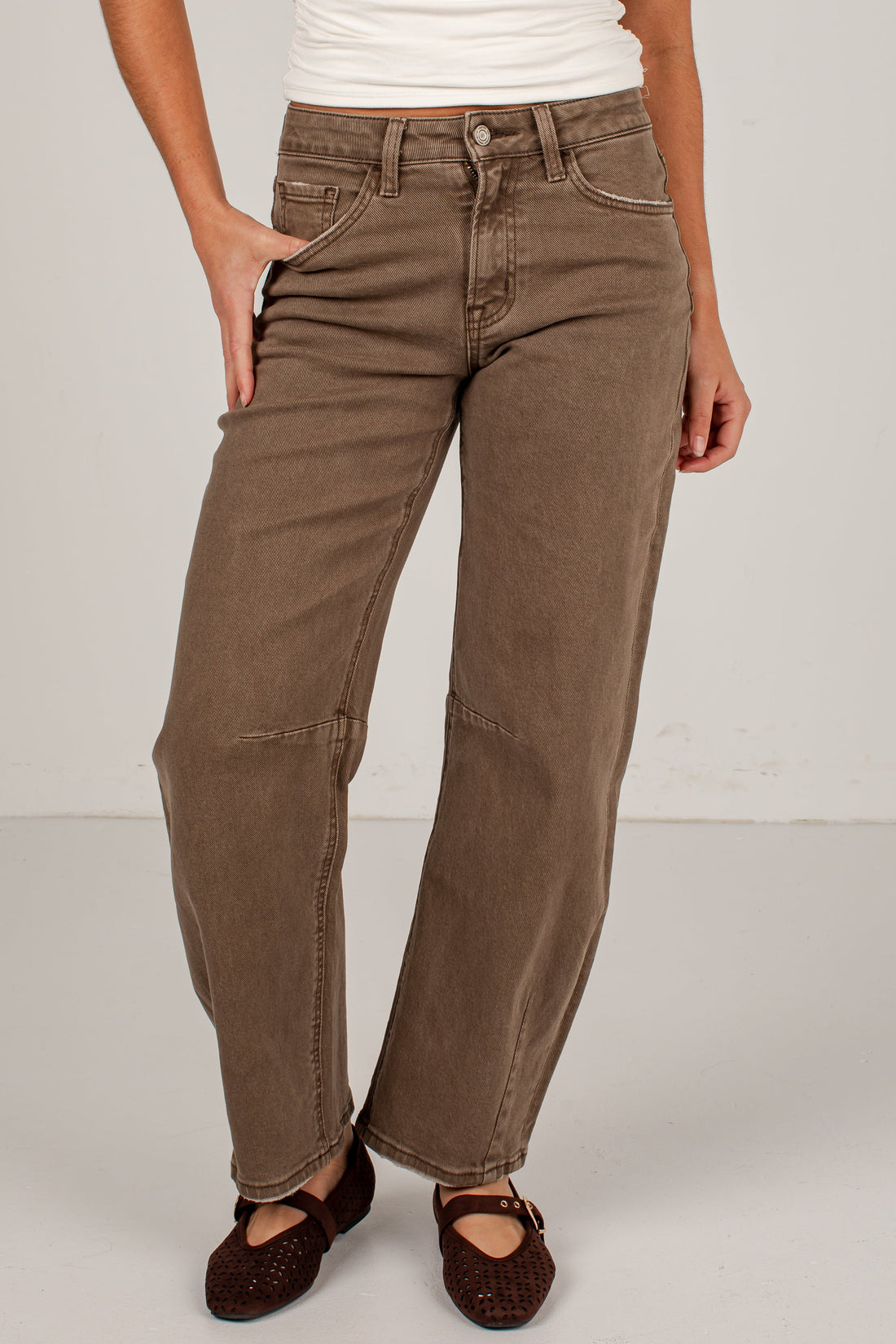 A person wearing brown wide-leg jeans with a white ribbed tank top and dark brown flat shoes, standing against a light gray studio background.