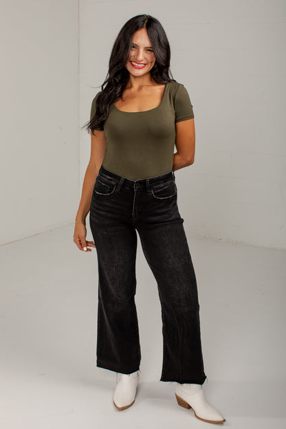 A woman modeling a fitted short-sleeve scoop-neck top in olive green, paired with black wide-leg jeans and white ankle boots, standing against a plain light gray studio background.