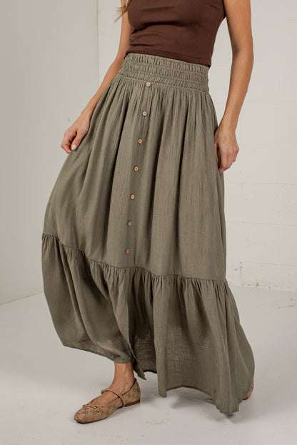 A woman wearing a high-waisted, tiered maxi skirt in a muted olive green color. The skirt features a smocked waistband, a vertical row of brown buttons down the front, and a flared, voluminous silhouette.