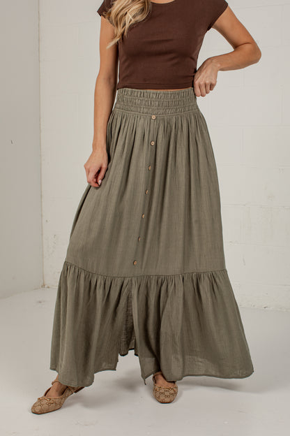 A woman wearing a long, tiered olive green skirt with a button-down front and a brown ribbed top, standing against a white studio wall.