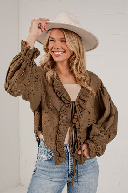 A woman wearing a brown eyelet lace cropped jacket with ruffle trim, a beige top, blue jeans, and a wide-brimmed beige hat, posing against a white brick wall.