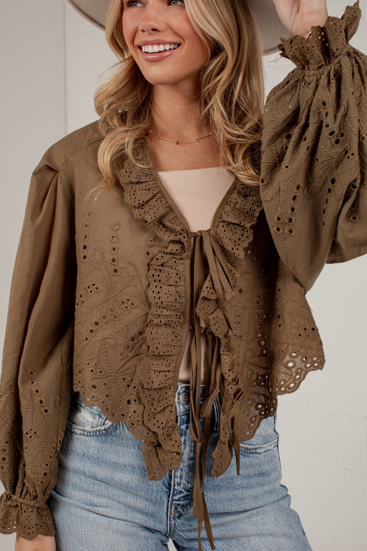 A woman wearing a brown eyelet lace cropped blouse with ruffles and tie-front detailing, paired with a beige top and blue jeans.