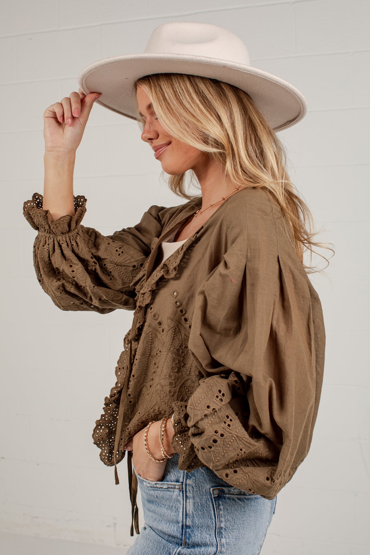 A woman wearing a brown eyelet blouse with ruffled trim, blue jeans, and a wide-brimmed cream hat, posing against a white paneled wall.