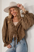 A woman wearing a brown eyelet lace long-sleeve blouse with ruffles and tie details, paired with a beige wide-brim hat and light blue jeans, posing against a white brick wall.