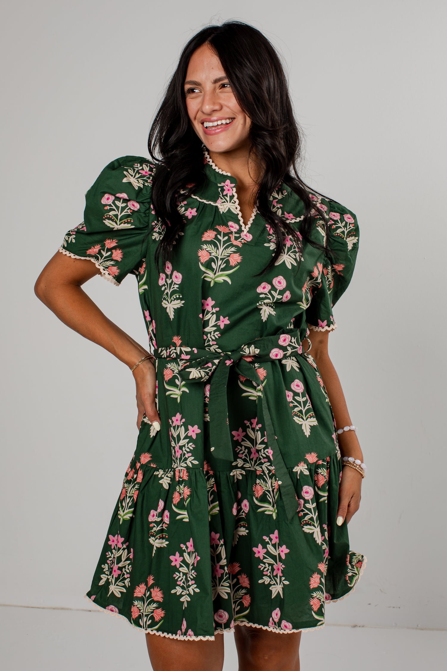 A woman modeling a dark green short-sleeve mini dress with a pink and white floral print, featuring puff sleeves, a collar, and a matching fabric tie belt, set against a plain light gray background.