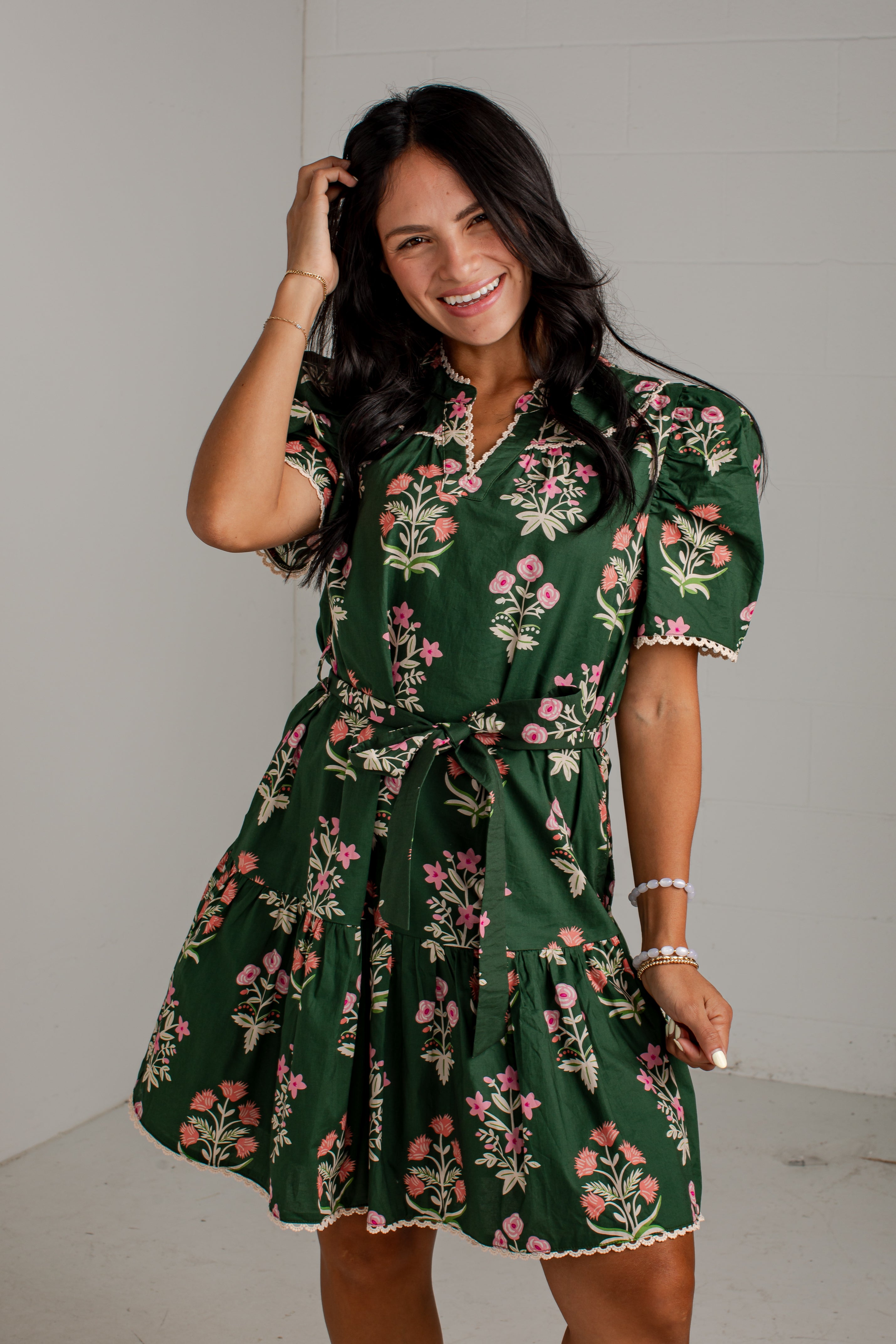 A woman modeling a dark green short-sleeve floral dress with pink and white flowers, a tie waist, and ruffled trim, standing against a light gray wall.