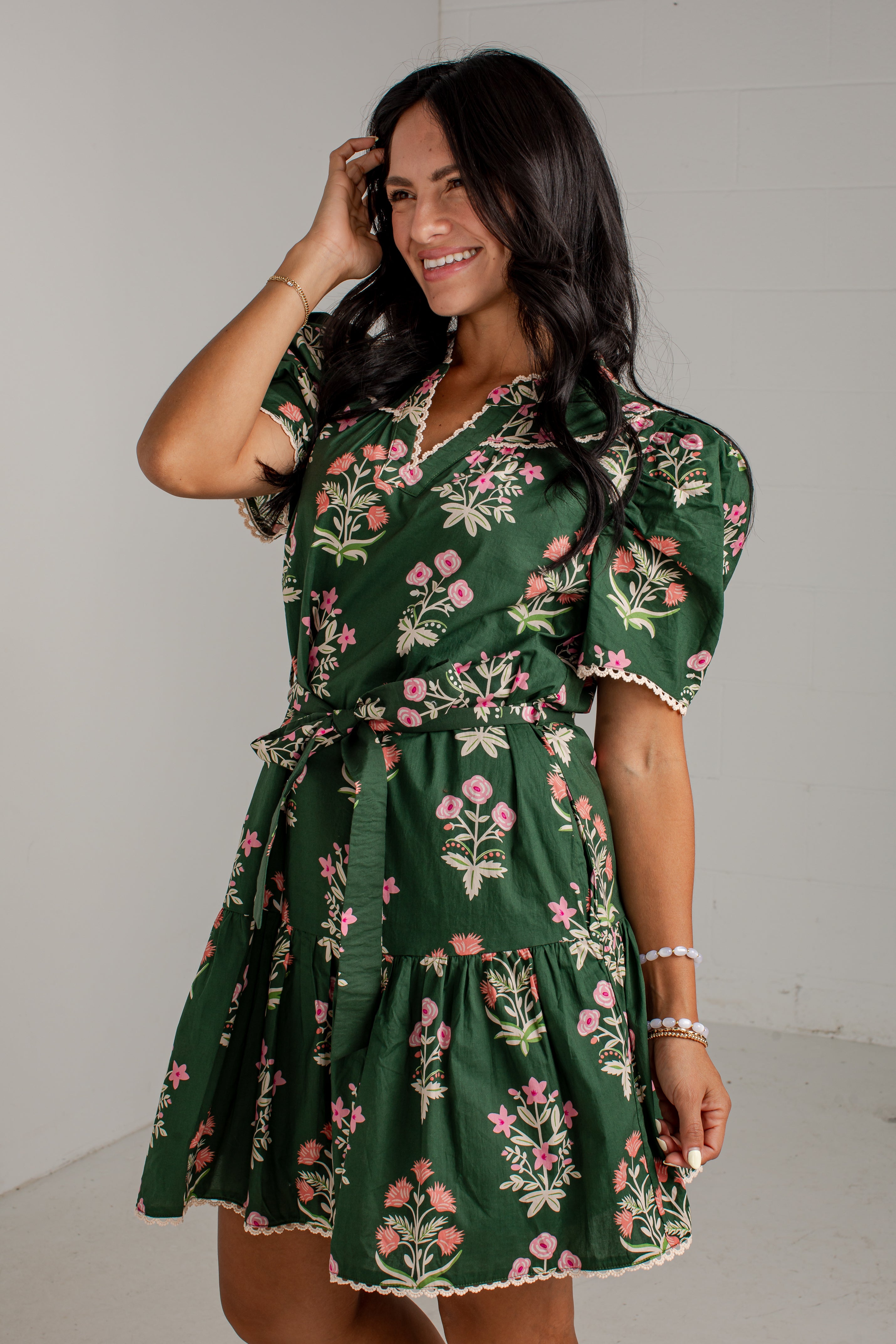 A woman modeling a dark green short-sleeve wrap dress with a pink and white floral print, featuring puff sleeves, a tie waist, and a tiered skirt.