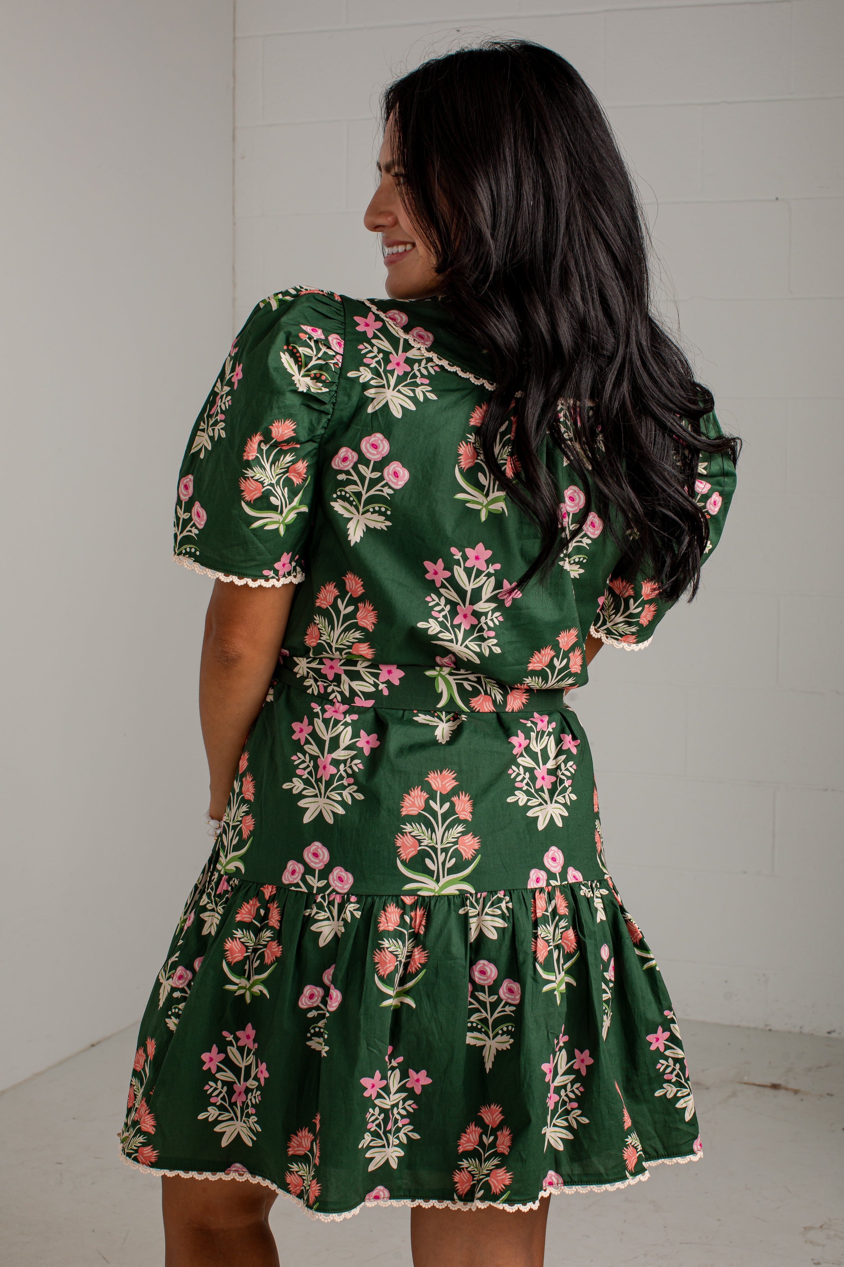 A woman wearing a dark green short-sleeve dress with a pink and white floral print, shown from the back against a white wall.