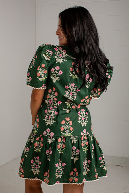 A woman wearing a dark green short-sleeve dress with a pink and white floral print, shown from the back against a white wall.