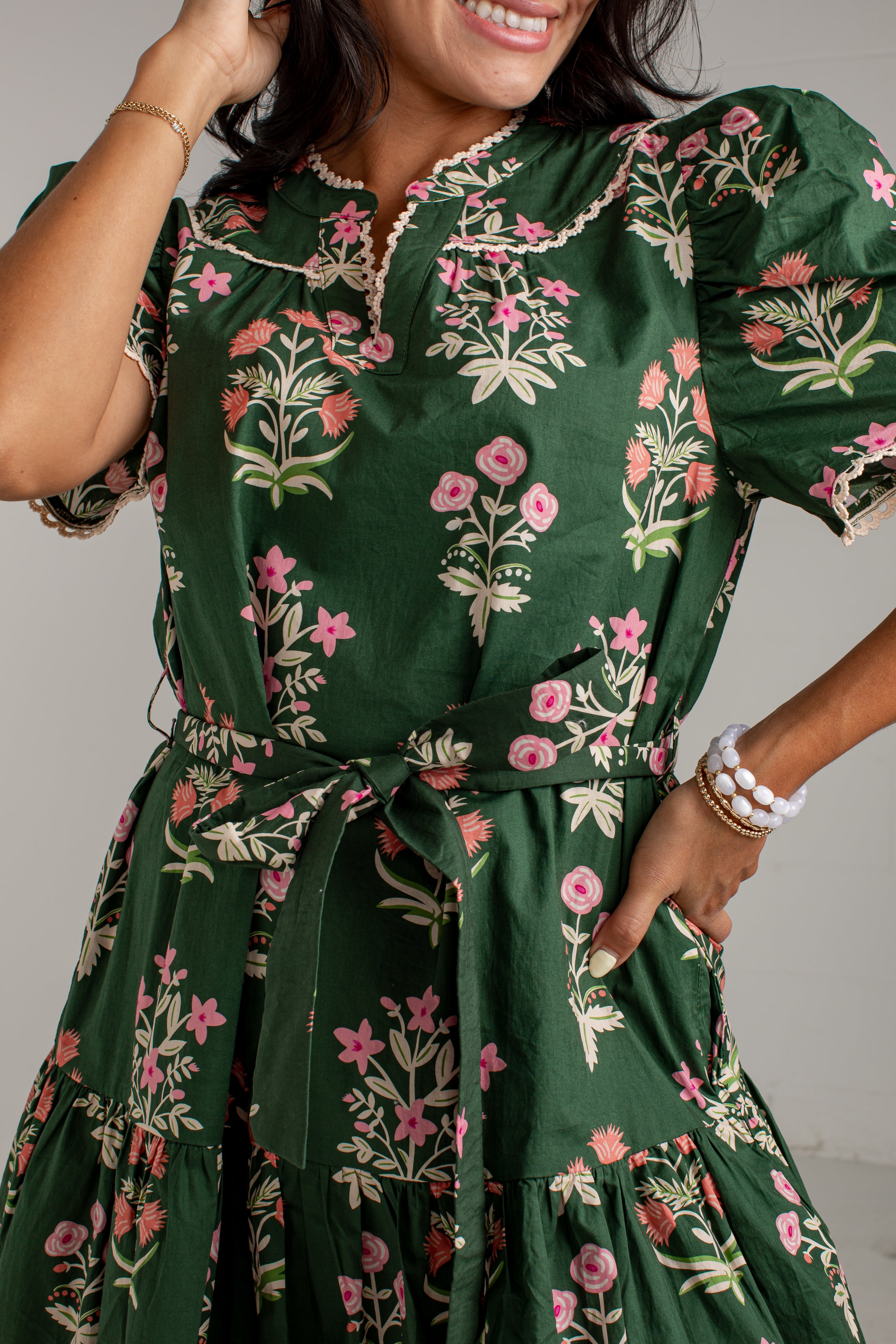 A woman wearing a dark green short-sleeve dress with a pink and beige floral print, featuring a tie waist and ruffled collar.