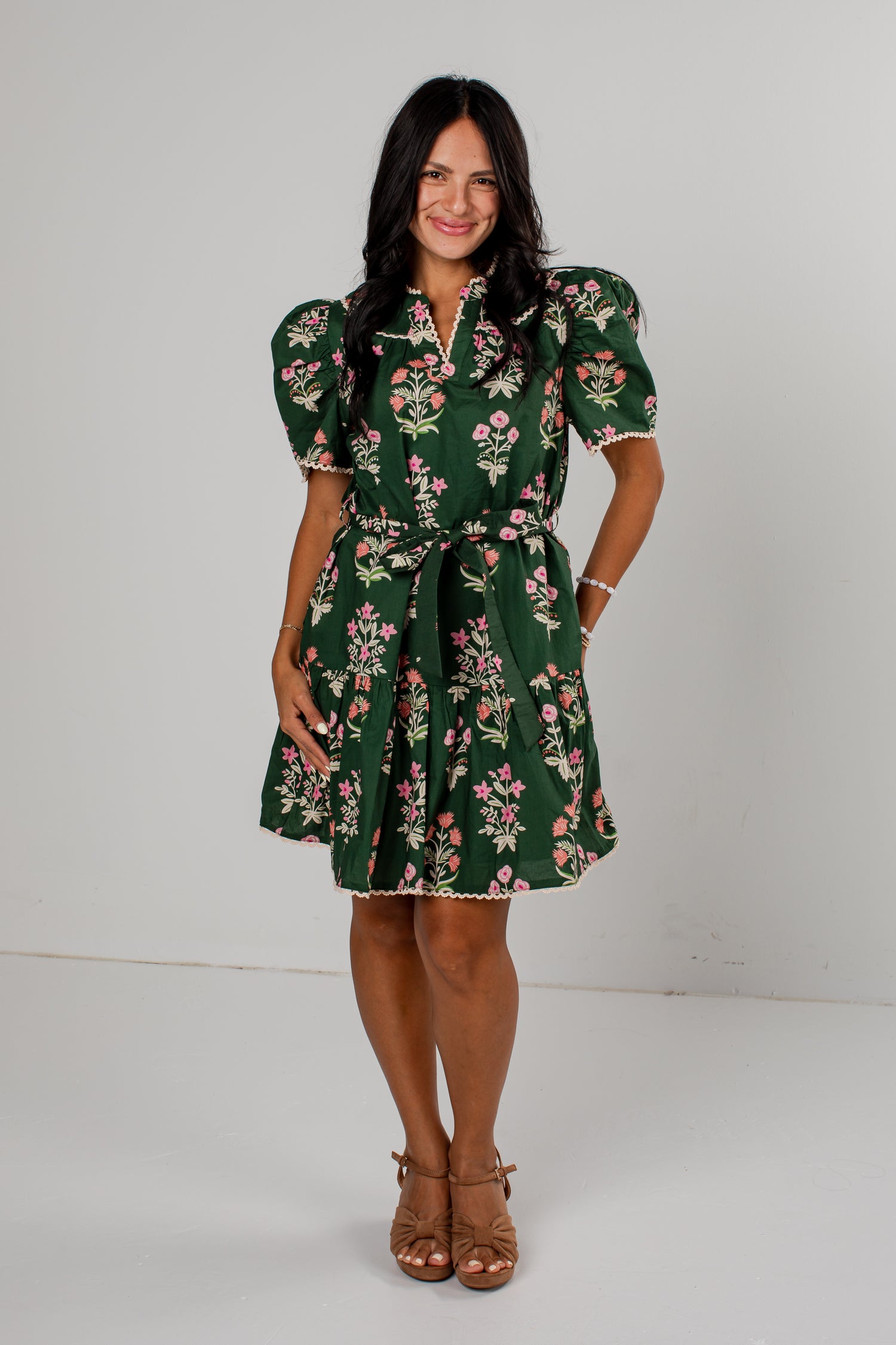A woman modeling a dark green short-sleeve mini dress with a pink and white floral print, puff sleeves, and a matching tie belt, standing against a plain light gray studio background.
