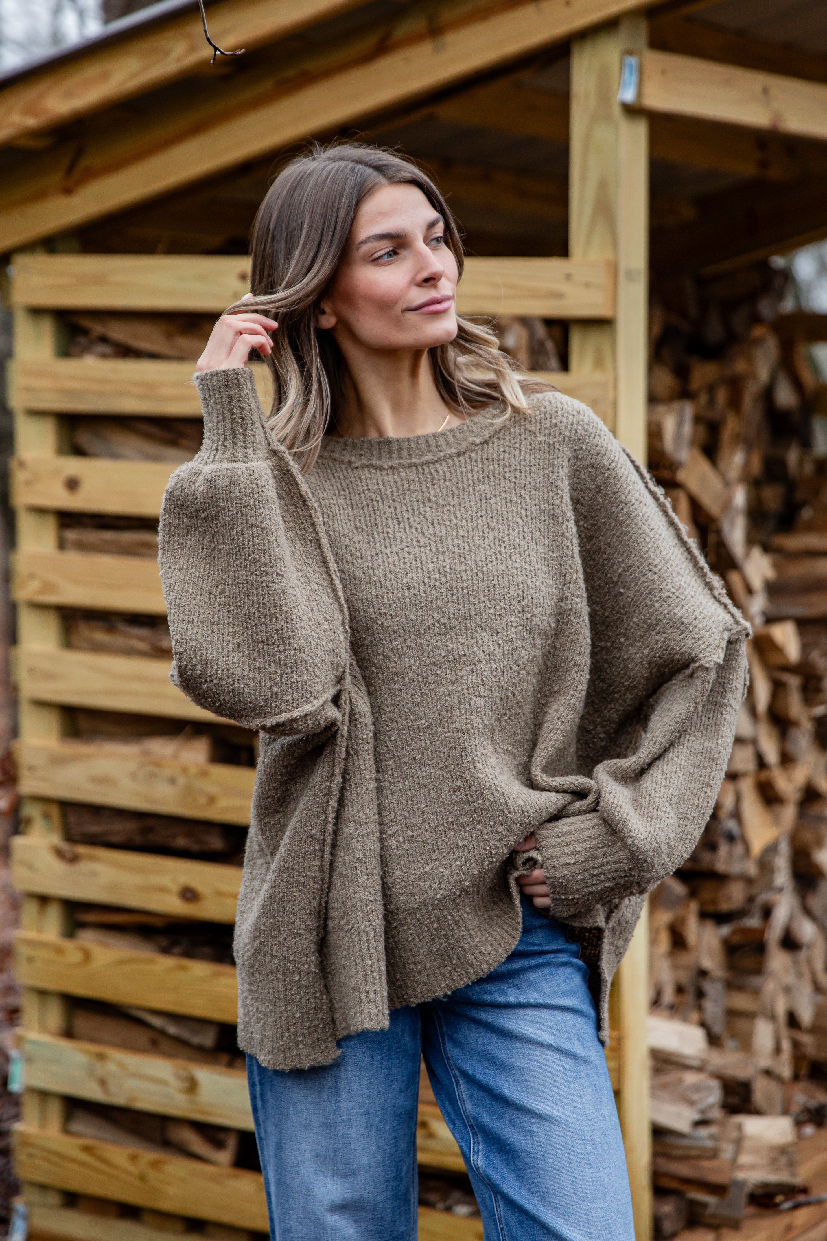 Woman wearing a brown poncho standing in front of a wooden structure with stacked firewood.