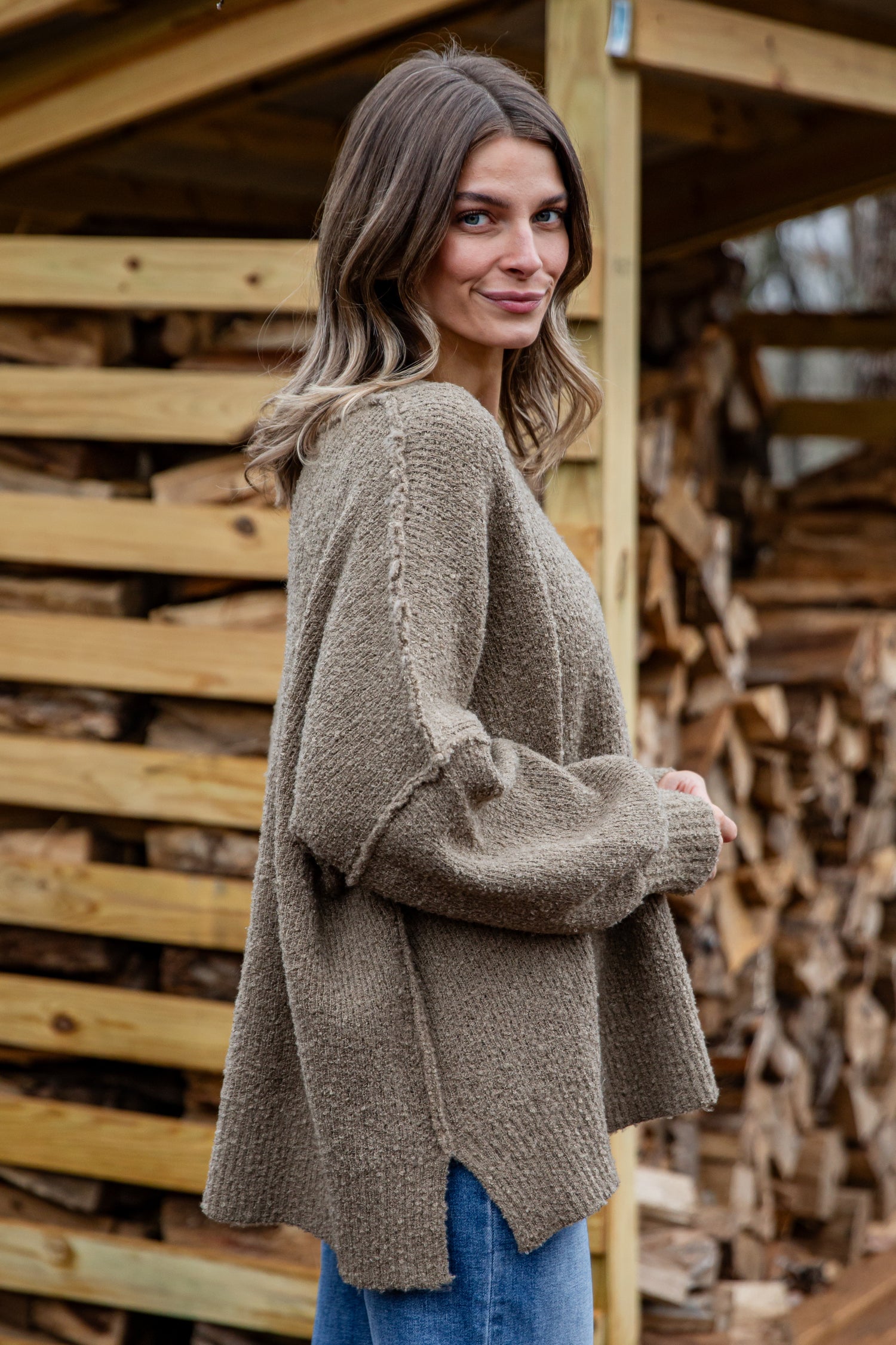 Woman wearing a brown sweater standing in front of stacked firewood.