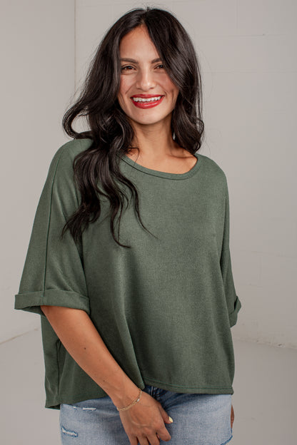 A woman with long dark hair wearing an olive green short-sleeve top and light blue jeans, standing against a plain light gray wall.